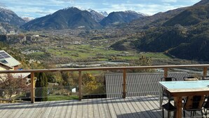 Une terrasse en bois avec une table et des chaises, offrant une vue sur une vallée, des montagnes et une ville.