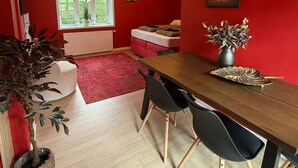 A room with a red wall, a wooden dining table, black chairs, a red sofa, a window with a view of greenery, and a ceiling-mounted smoke detector. - Four Seasons - Crimson Autumn Suite (Aalborg)