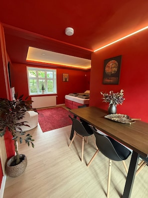 A room with a red wall, a wooden dining table, black chairs, a red sofa, a window with a view of greenery, and a ceiling-mounted smoke detector.