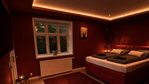 A bedroom with a bed, a window, a radiator, a red rug, and a small table with a lamp. - Four Seasons - Crimson Autumn Suite (Aalborg)