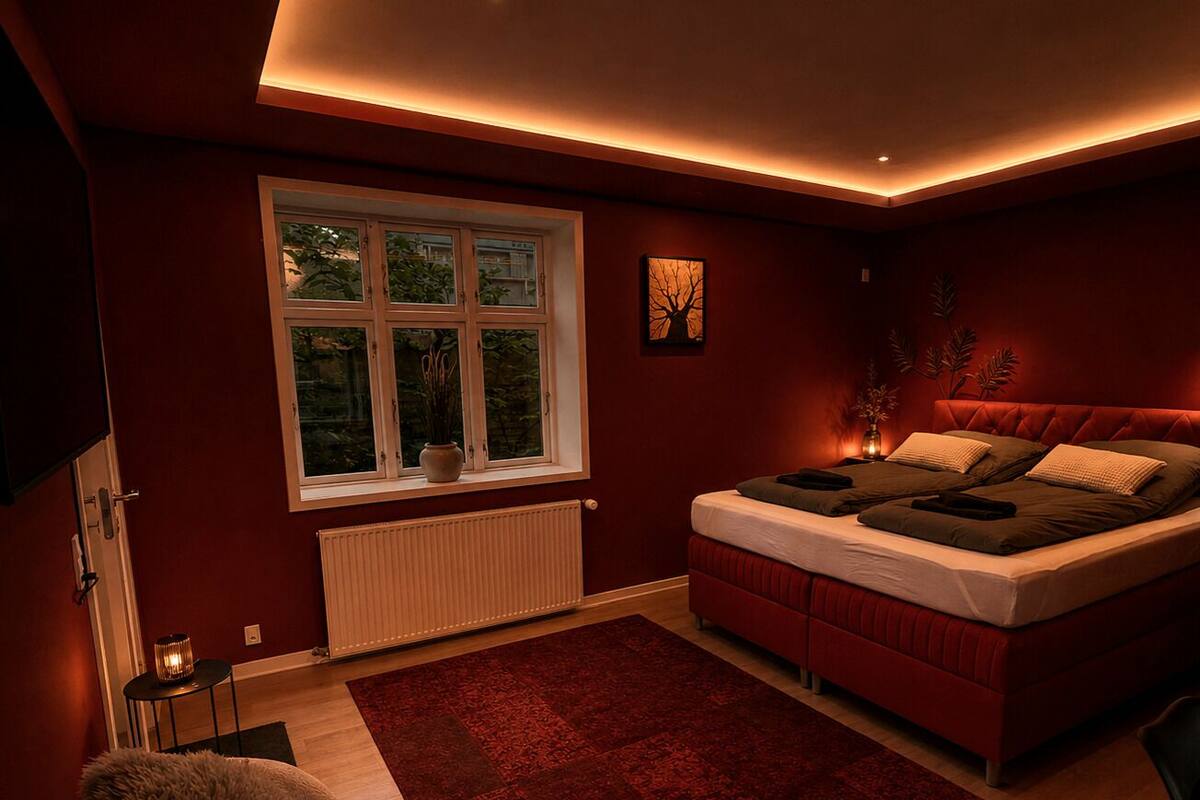 A bedroom with a bed, a window, a radiator, a red rug, and a small table with a lamp.