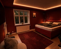 Un dormitorio con cama, ventana, radiador, alfombra roja y una pequeña mesa con lámpara.