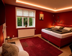 A bedroom with a red color scheme, a large window with blinds, a bed with multiple pillows, a radiator, and a red rug.
