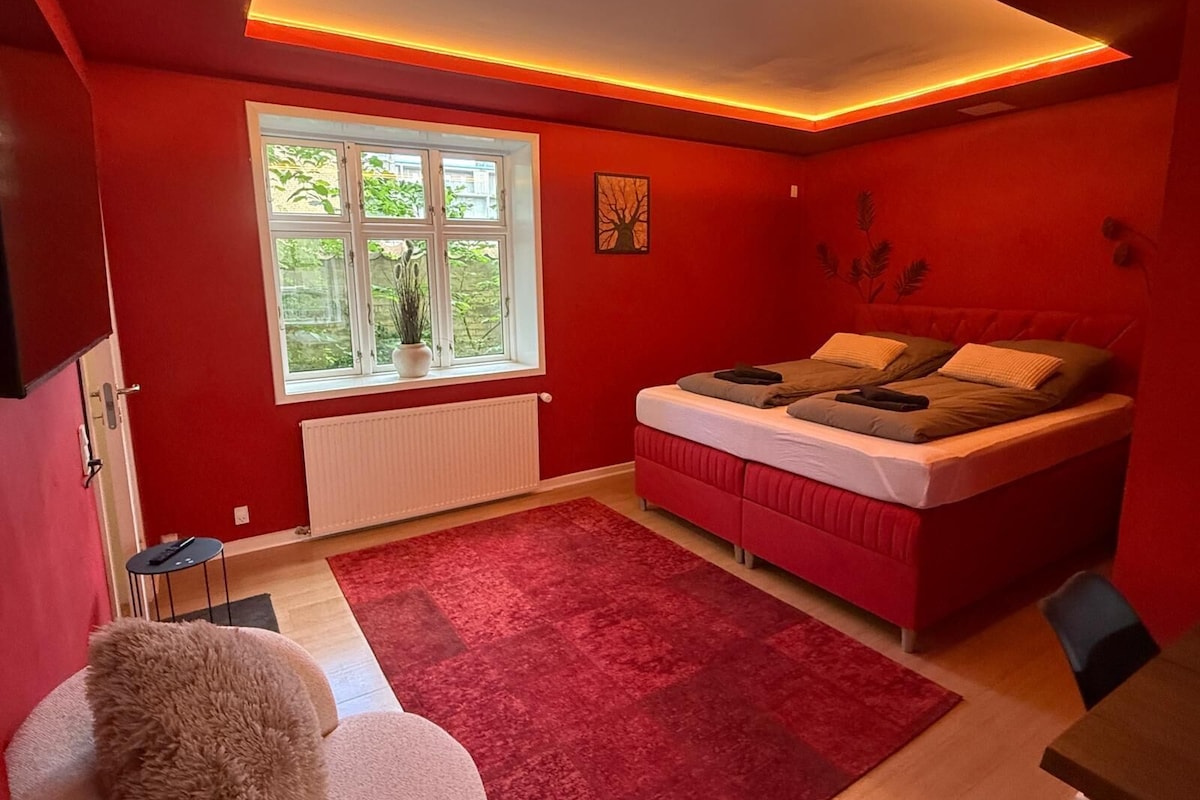 A bedroom with a red bed, a window, a radiator, and a red carpet.