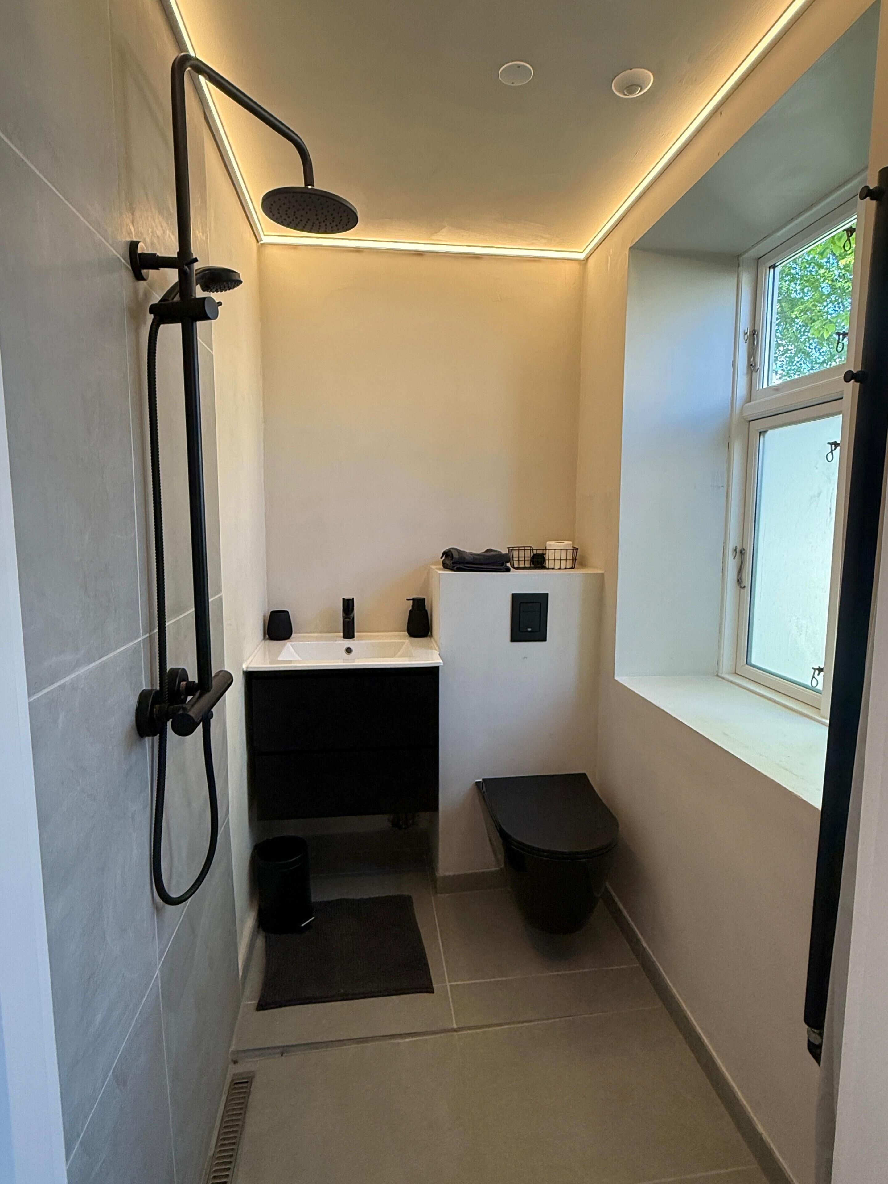 A small bathroom with a shower, sink, and toilet, featuring a window and a black mat.