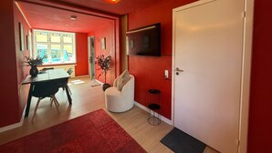 A modern living room with a red wall, a white door, a flat-screen TV, a dining area with a table and chairs, and a large window.
