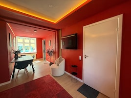 Una sala de estar moderna con una pared roja, una puerta blanca, un televisor de pantalla plana, una zona de comedor con mesa y sillas, y un ventanal grande.