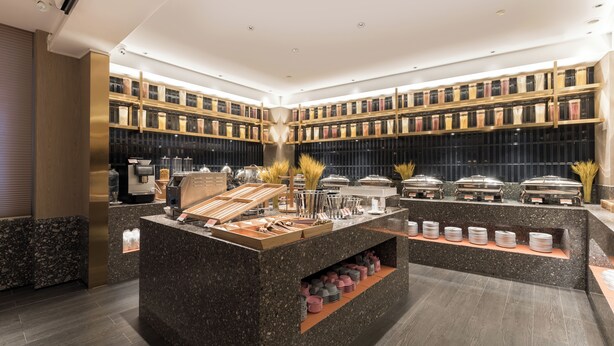 A modern hotel lobby with a central buffet area, multiple shelves of books, and a well-lit ceiling. - Paco Hotel Canton Tower Pazhou (Guangzhou)