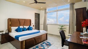 Quarto de hotel com uma cama grande, uma escrivaninha, uma cadeira, um ventilador de teto e uma janela com vista para o céu.
