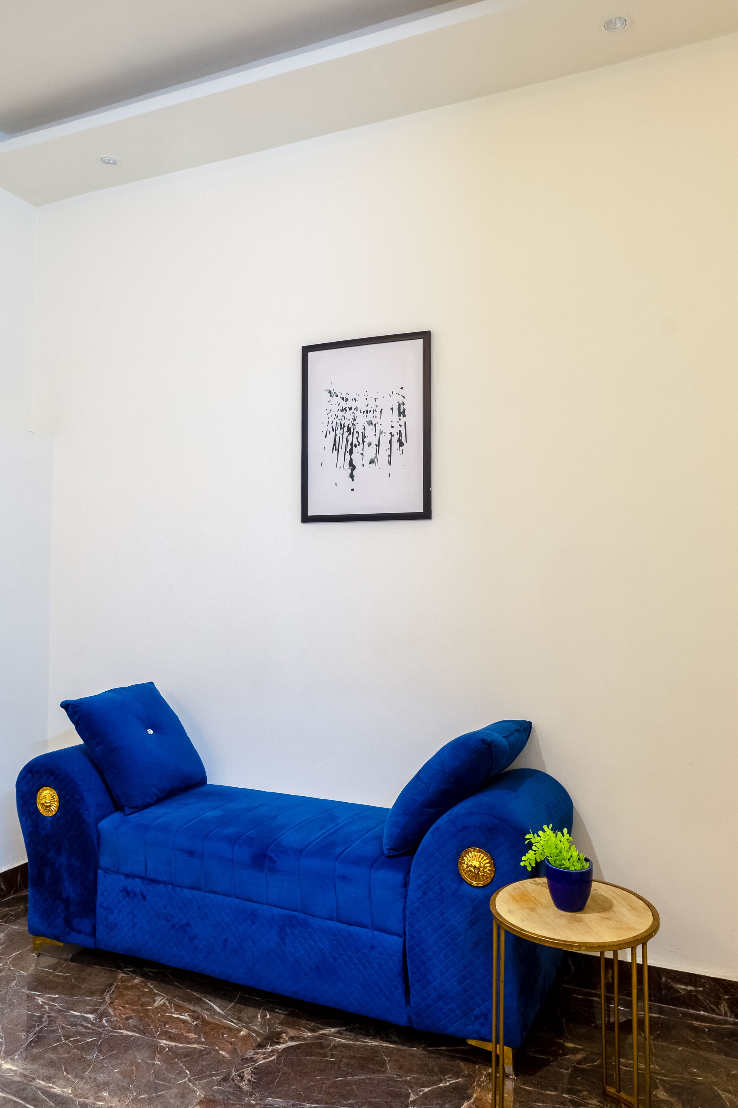 Un canapé bleu agrémenté de détails dorés, une petite table ronde sur laquelle est posée une plante, et une œuvre d’art abstraite encadrée accrochée au mur.