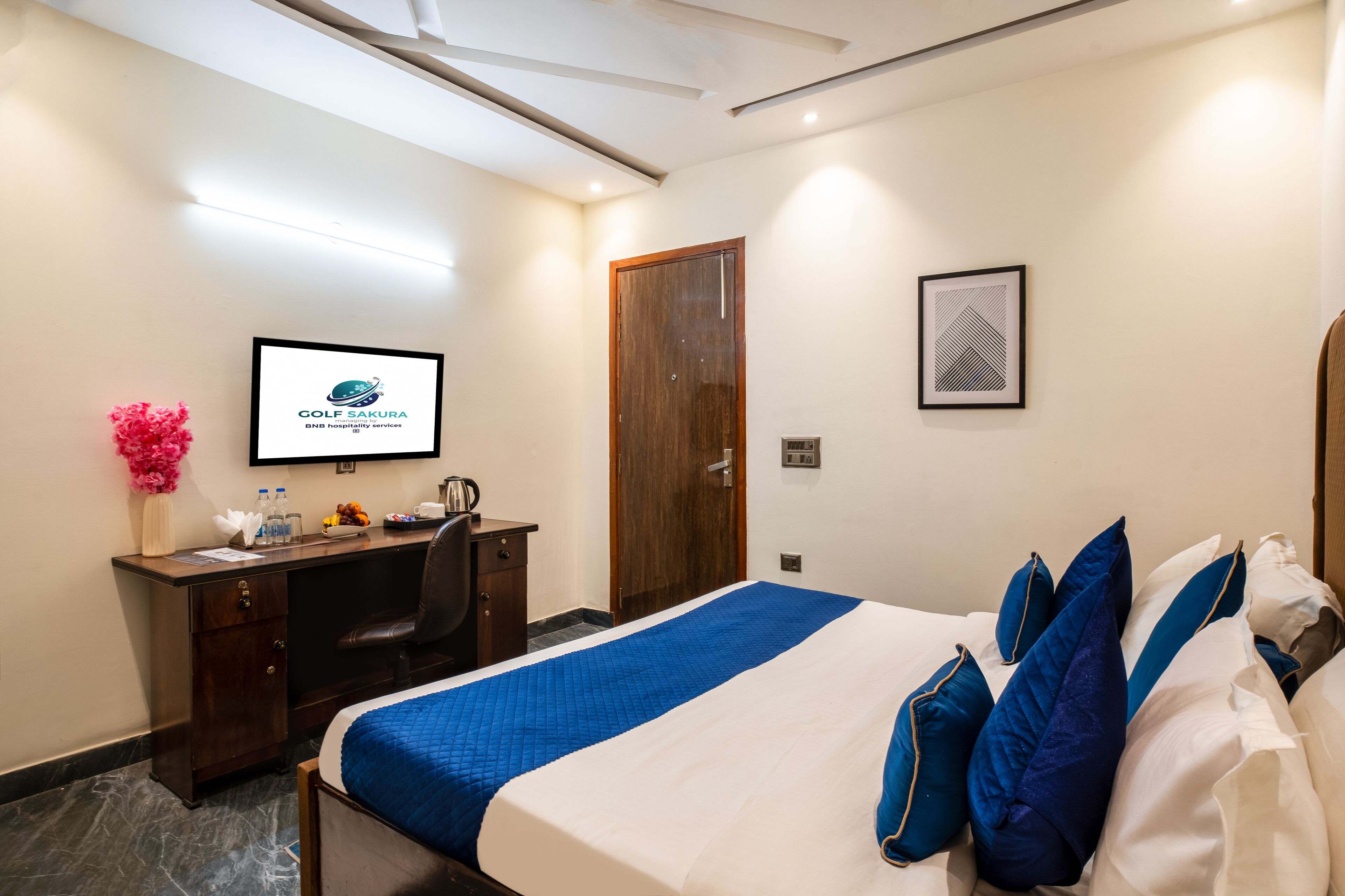 A hotel room with a bed, a desk, a chair, a TV, a door, and a framed picture on the wall.