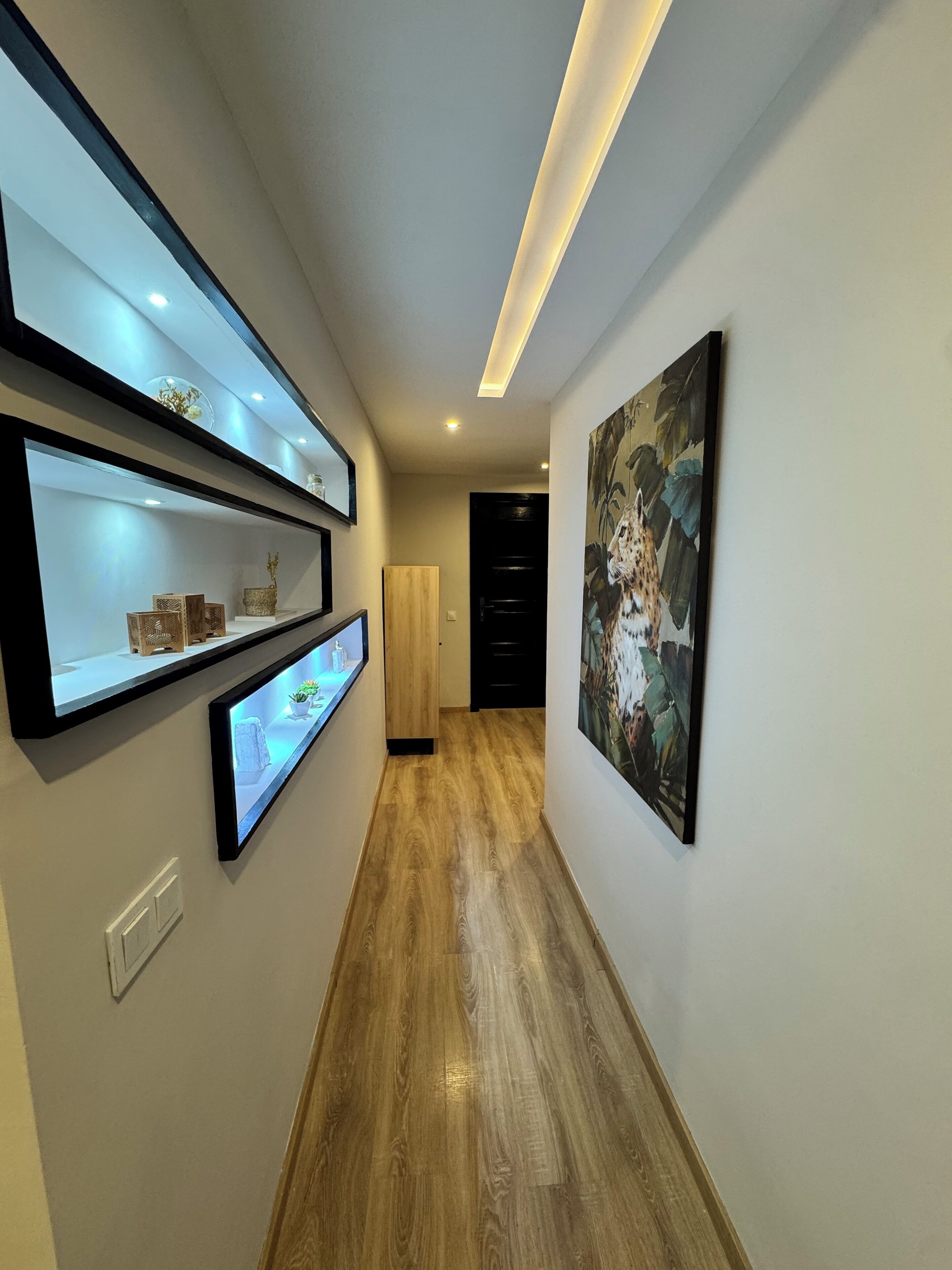 Een gang met houten vloer, wandplanken met decoratieve objecten, inbouwverlichting en een gekaderd schilderij aan de muur.