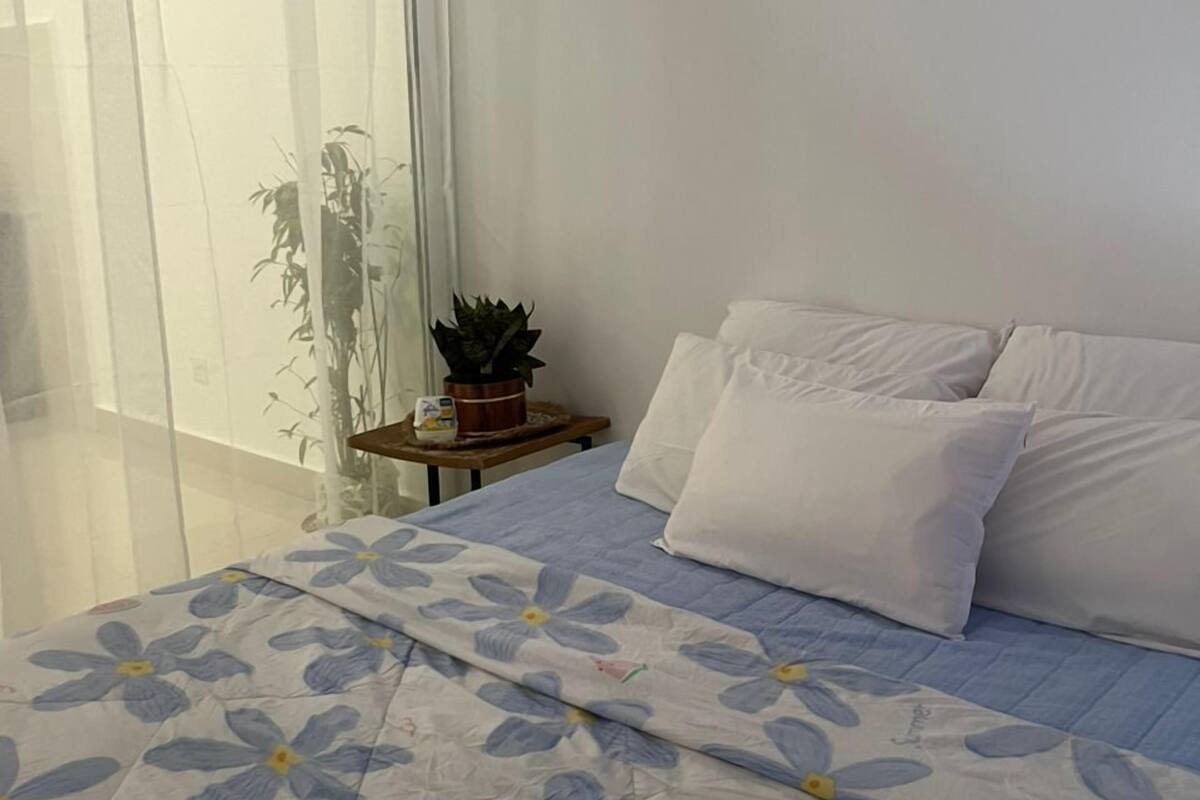 Un dormitorio con cama, almohadas blancas, mesita de noche con una planta y ventana con cortinas de encaje.