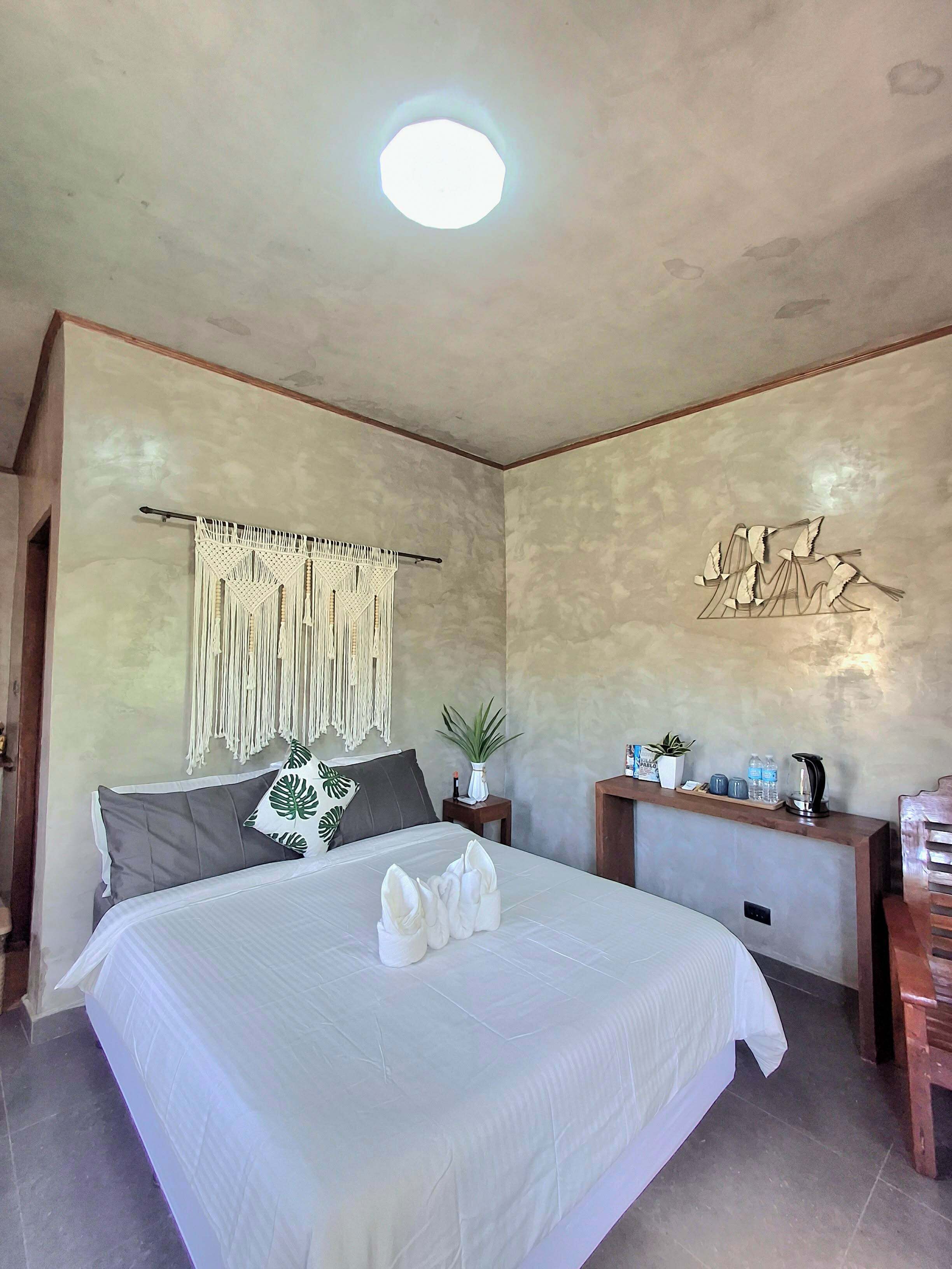 A bedroom with a large bed, a bedside table, a wall-mounted macrame decoration, a potted plant, and a wall art piece.