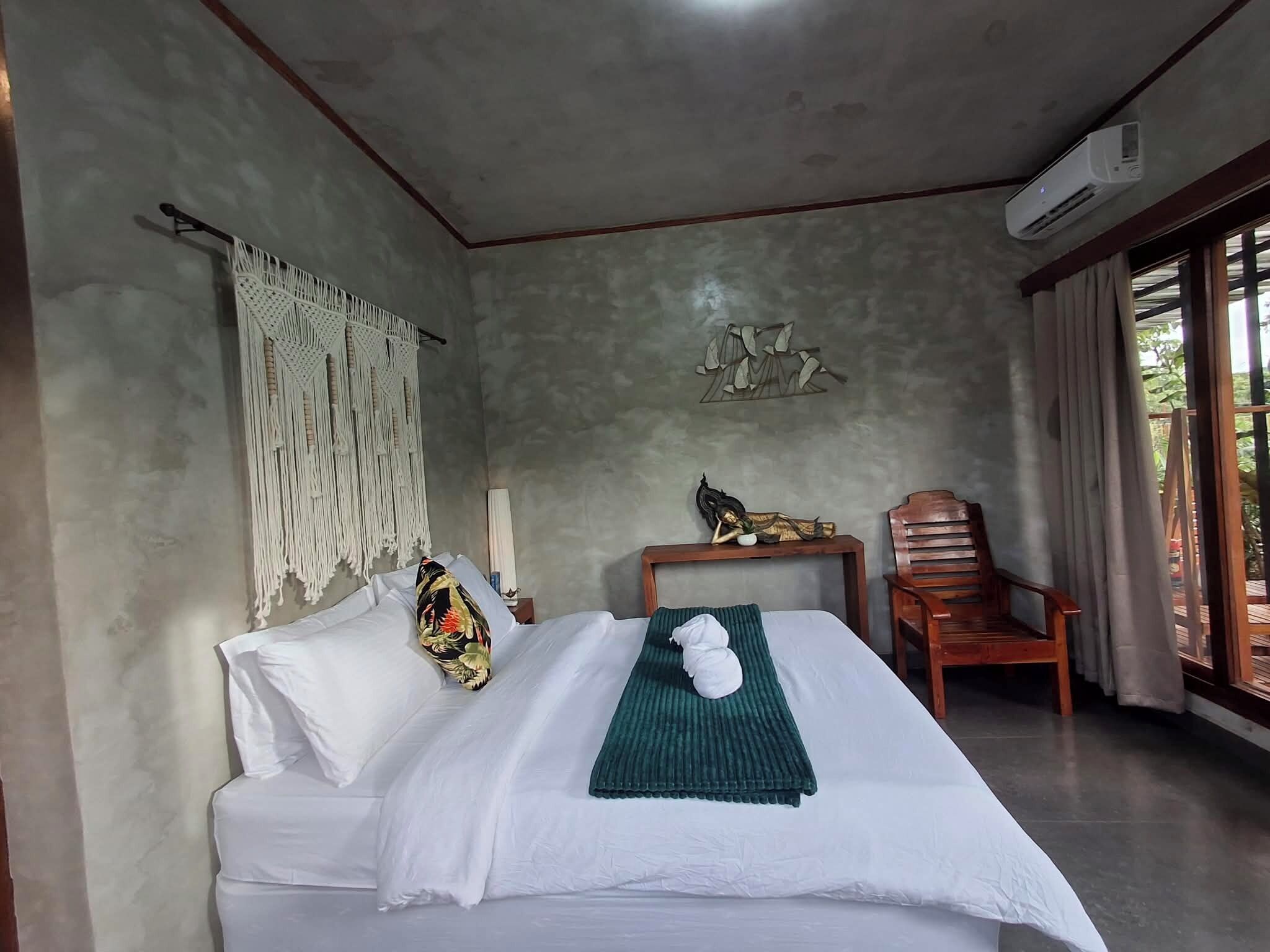 Een slaapkamer met een bed, een macramé wanddecoratie, een beeldje, een houten stoel en een airconditioning.