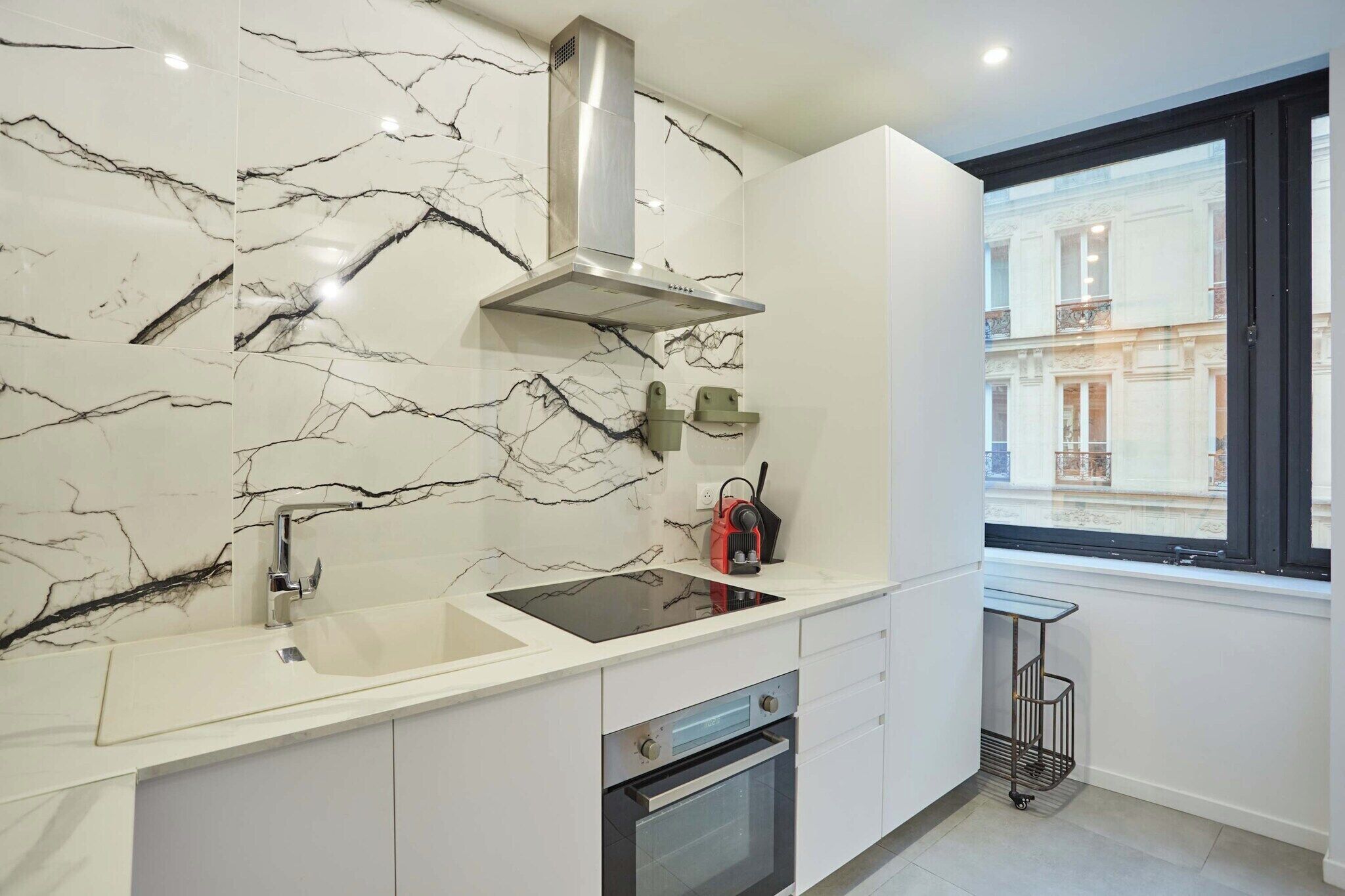 Een moderne keuken met een marmeren spatwand, een roestvrijstalen afzuigkap en een raam met uitzicht op een gebouw.