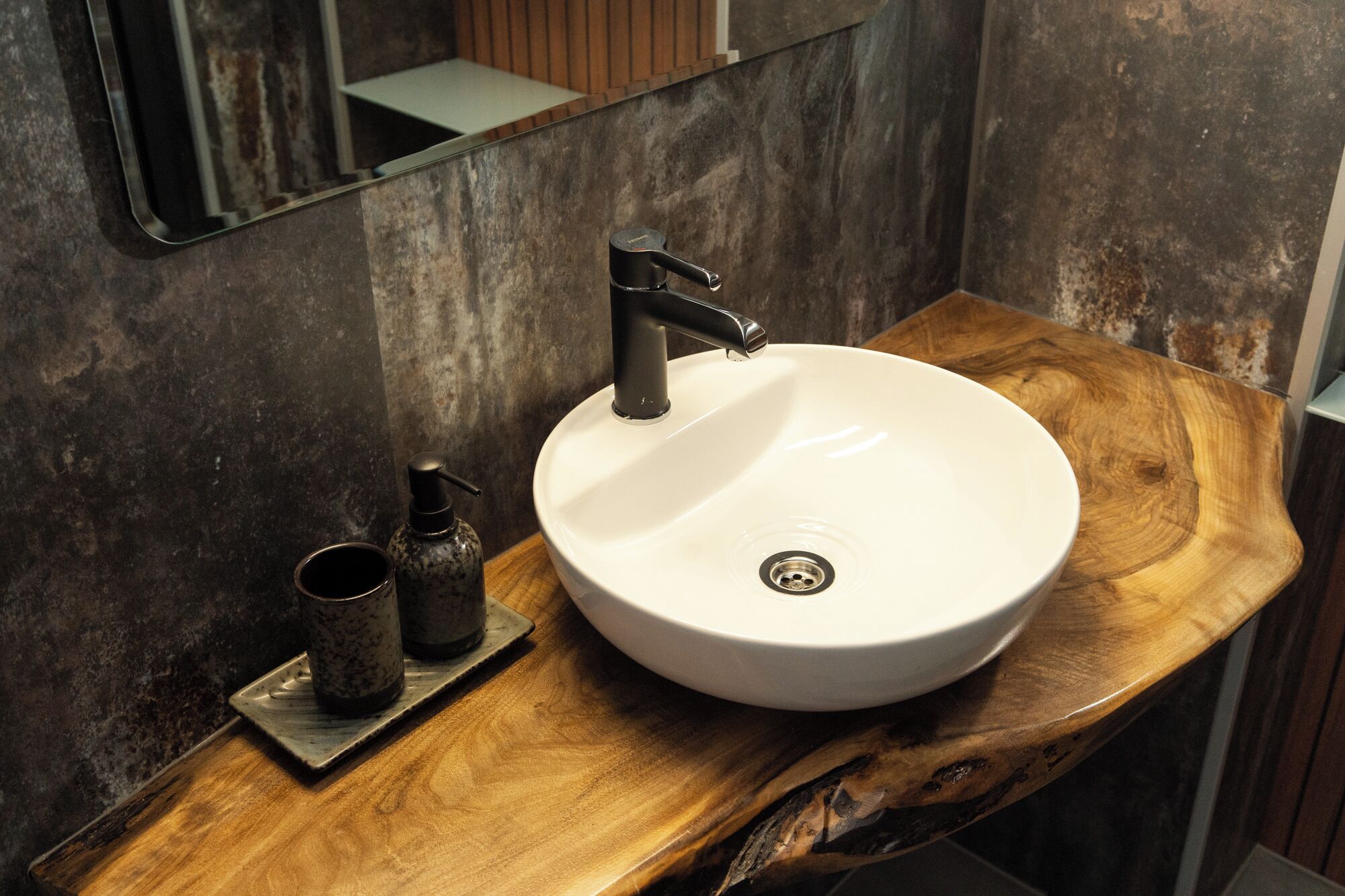 Un baño con un lavabo blanco y redondo, un grifo negro y una pequeña botella de jabón sobre una encimera de madera.