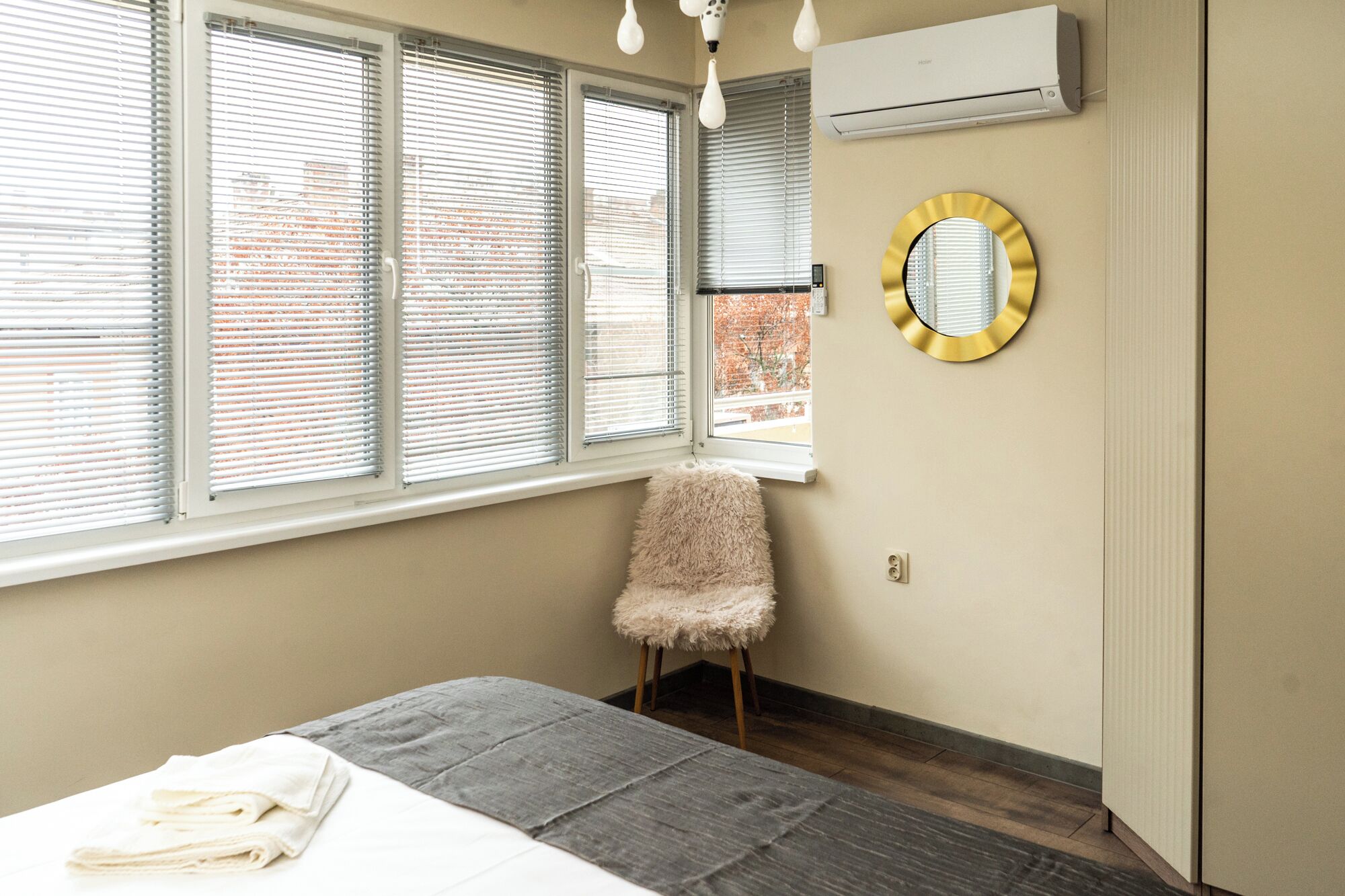 A bedroom with a bed, a fluffy chair, a round mirror, and an air conditioner.