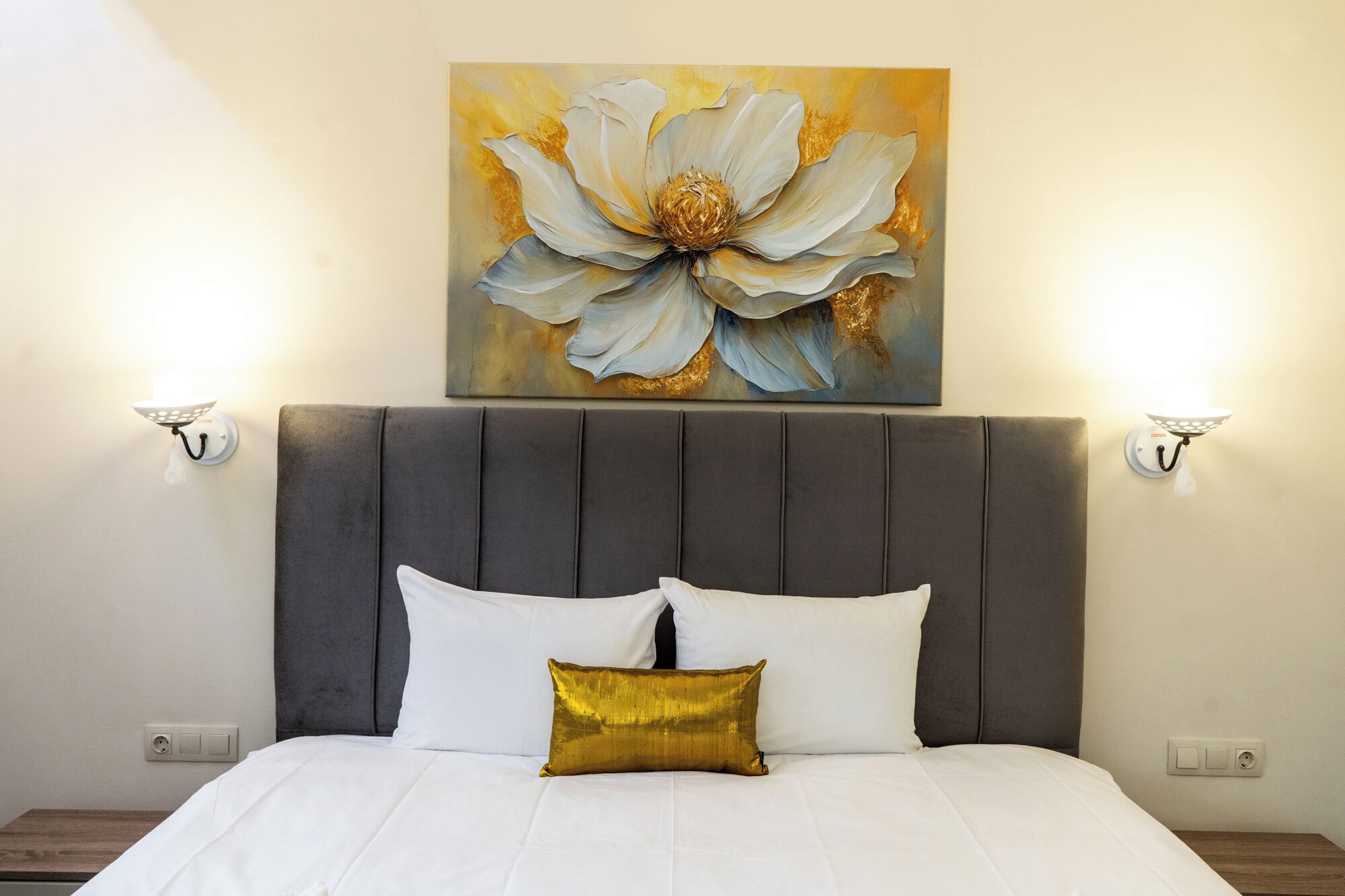 Ein Schlafzimmer mit einem großen Bett, einem grau gepolsterten Kopfteil und einem an der Wand befestigten Blumenbild.