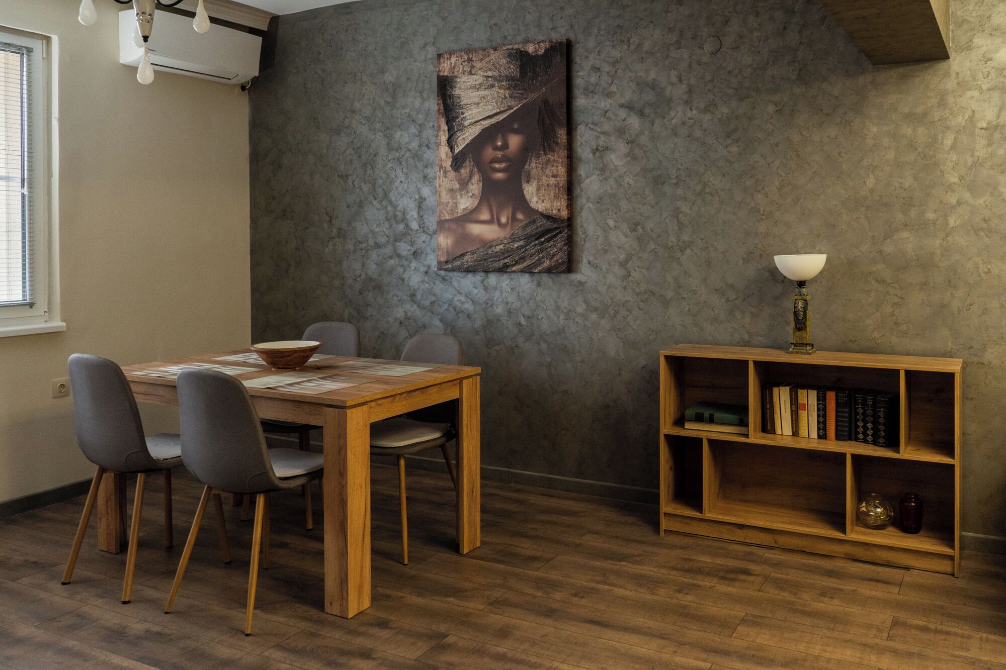 A dining area with a wooden table, grey chairs, a bookshelf, and a painting on the wall.