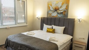 A neatly made bed with a grey headboard, white linens, and a gold accent pillow. There are two wall-mounted lights and a small wooden nightstand with drawers. - Apartment 8 (Lovech)