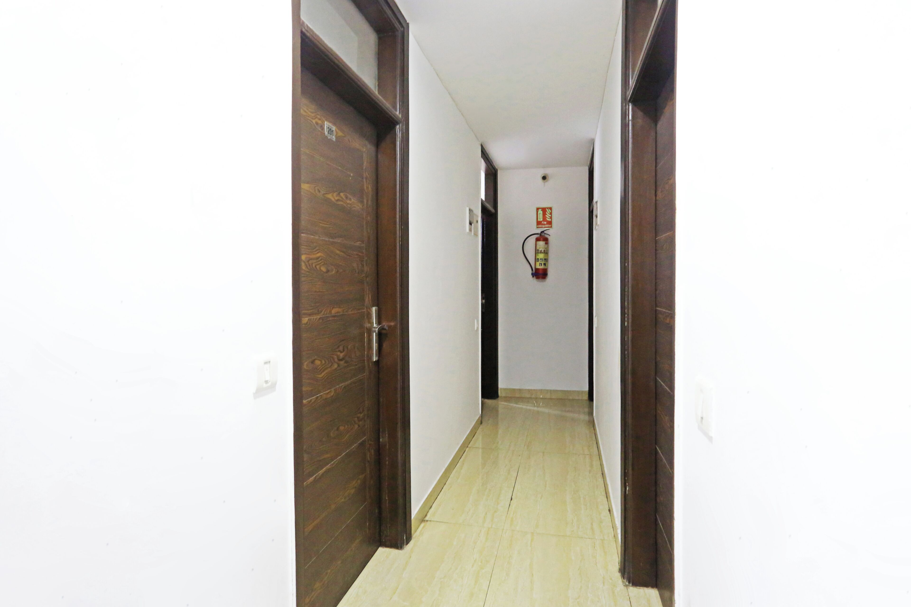 A narrow hallway with wooden doors on either side, a fire extinguisher mounted on the wall, and a tiled floor.
