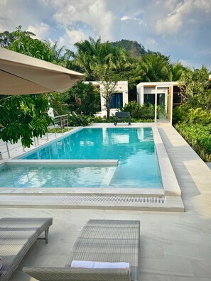 A rectangular swimming pool with a clear blue water surface, surrounded by a tiled deck. There are two lounge chairs with white cushions and a bench. The pool area is bordered by lush greenery and a mountain in the background. - Zana Villas (Krabi)