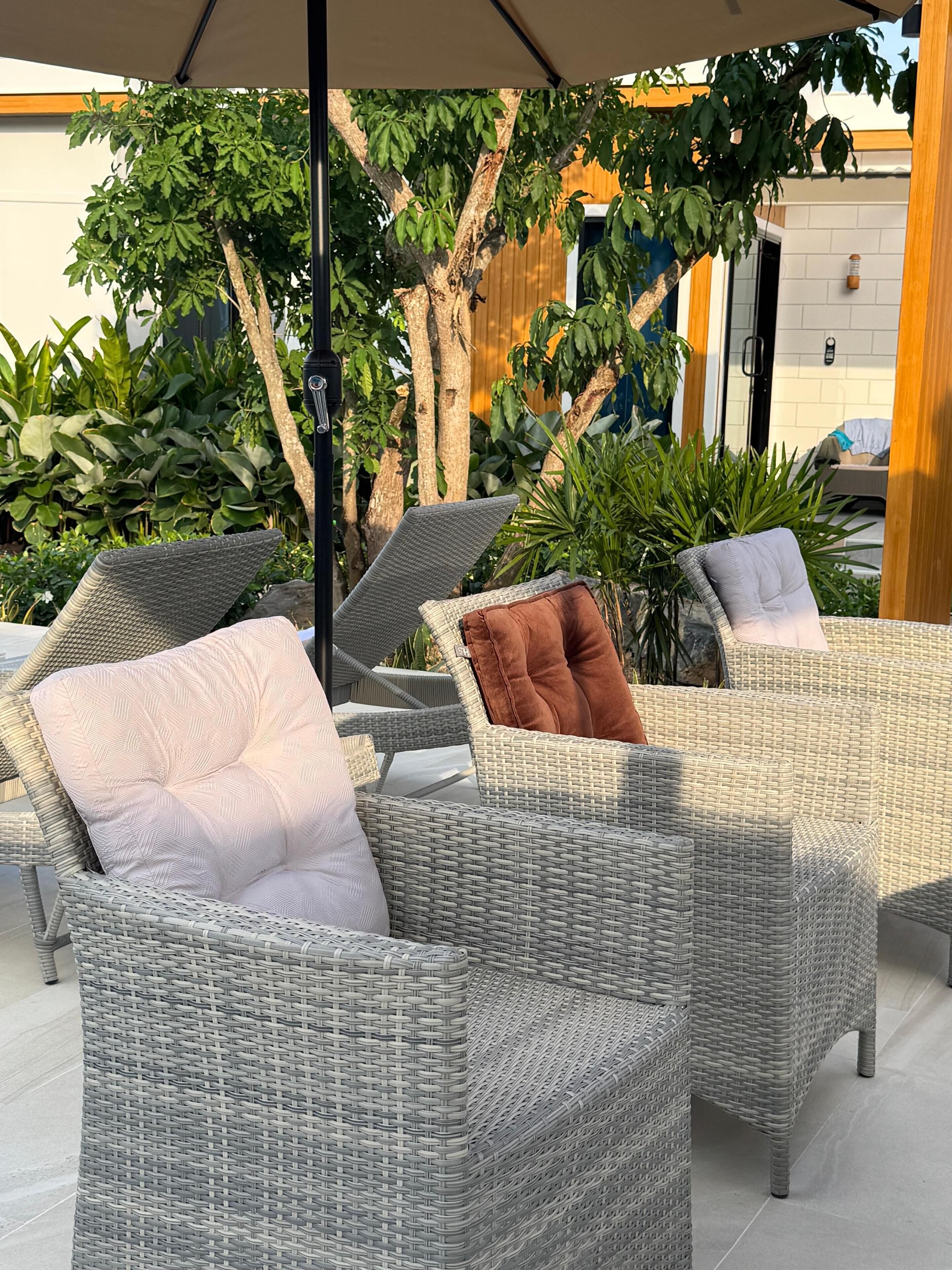 Een terras met rieten meubels en kussens, omgeven door groen en een gebouw op de achtergrond.