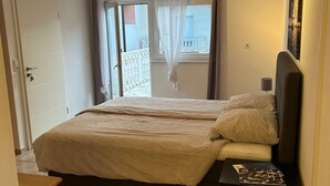A bedroom with a bed, a bedside table with a book, a window with curtains, and an air conditioning unit. - Cozy 2 room apartment with all amenities near krka park (Bilice)