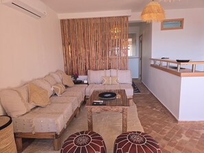 A living room with a sofa, ottomans, a coffee table, and a wooden partition. - Taghazout Ait Bihi Cosy short-term accommodation (Ait Bihi)