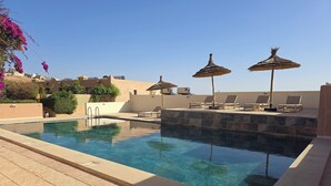 Una piscina con sillas de descanso y sombrillas, rodeada por un suelo de baldosas y un muro bajo.