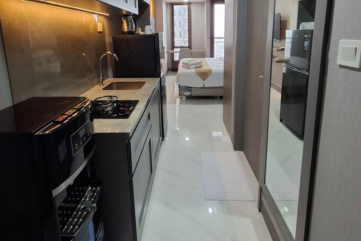 Cozinha moderna com fogão a gás, forno e micro-ondas. Há uma área de jantar com mesa e cadeiras. Uma cama é visível ao fundo.