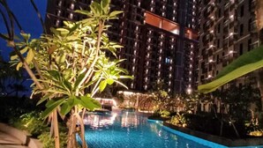 A nighttime view of a swimming pool surrounded by lush greenery and a high-rise building with illuminated windows. - Apartmen Pakuwon Bekasi by Ruang Singgah (Bekasi)