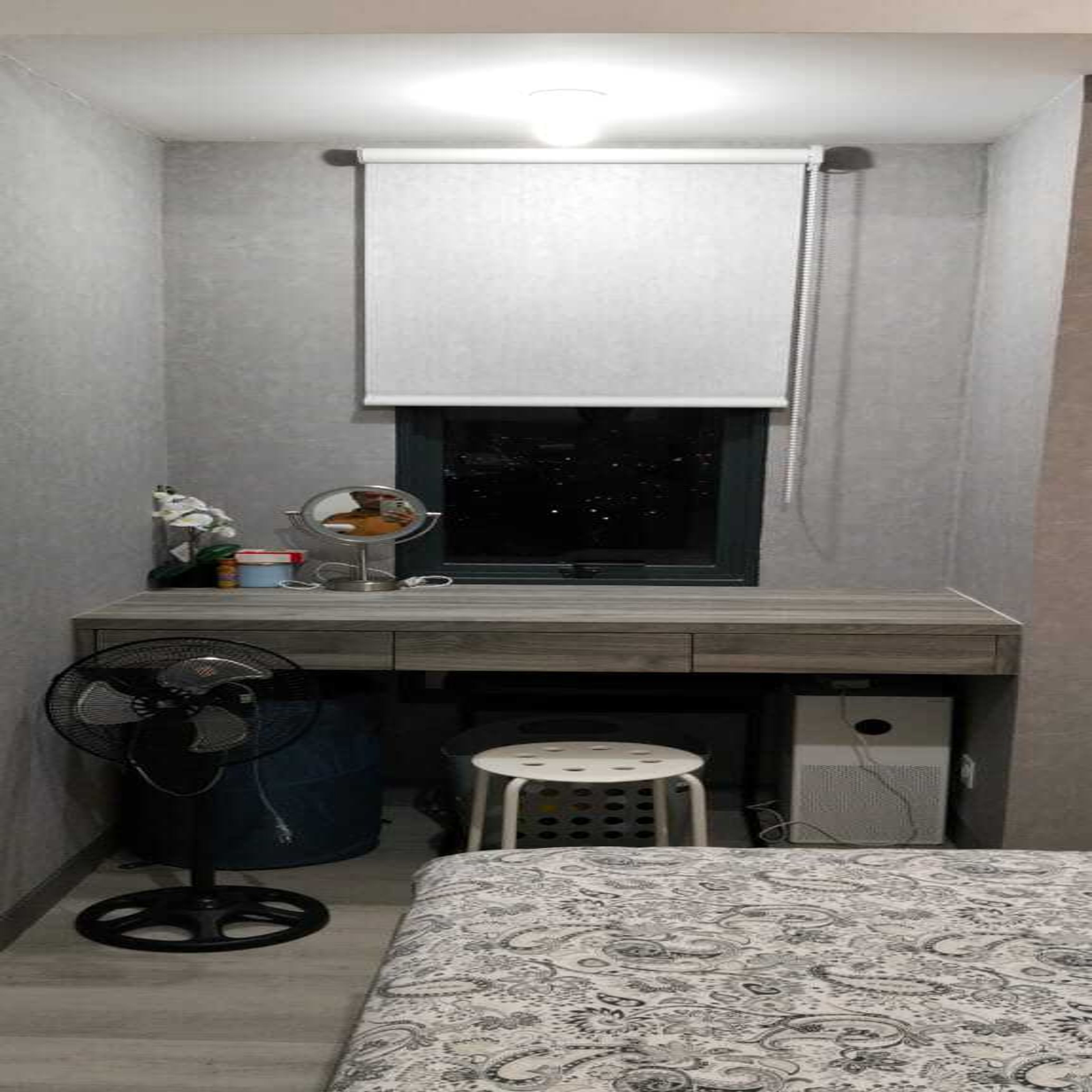 A small, dimly lit room with a desk, a chair, a small stool, a fan, and a window with a blind.