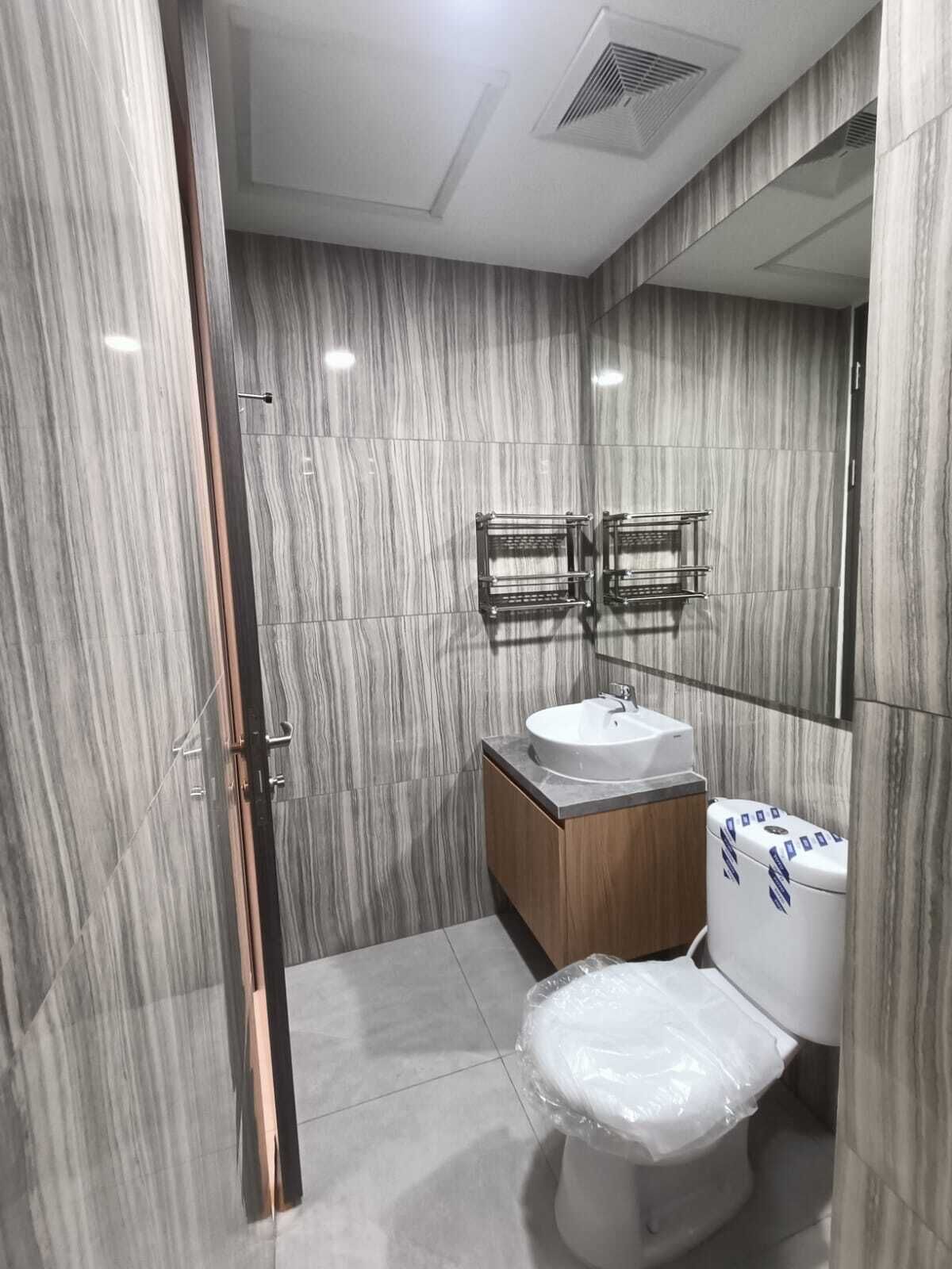 A modern bathroom with a marble wall, a wooden vanity, a white sink, and a toilet with a plastic cover.
