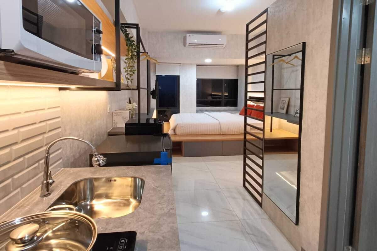 Cozinha compacta com micro-ondas, pia e fogão, que dá acesso a um quarto com cama e televisão.