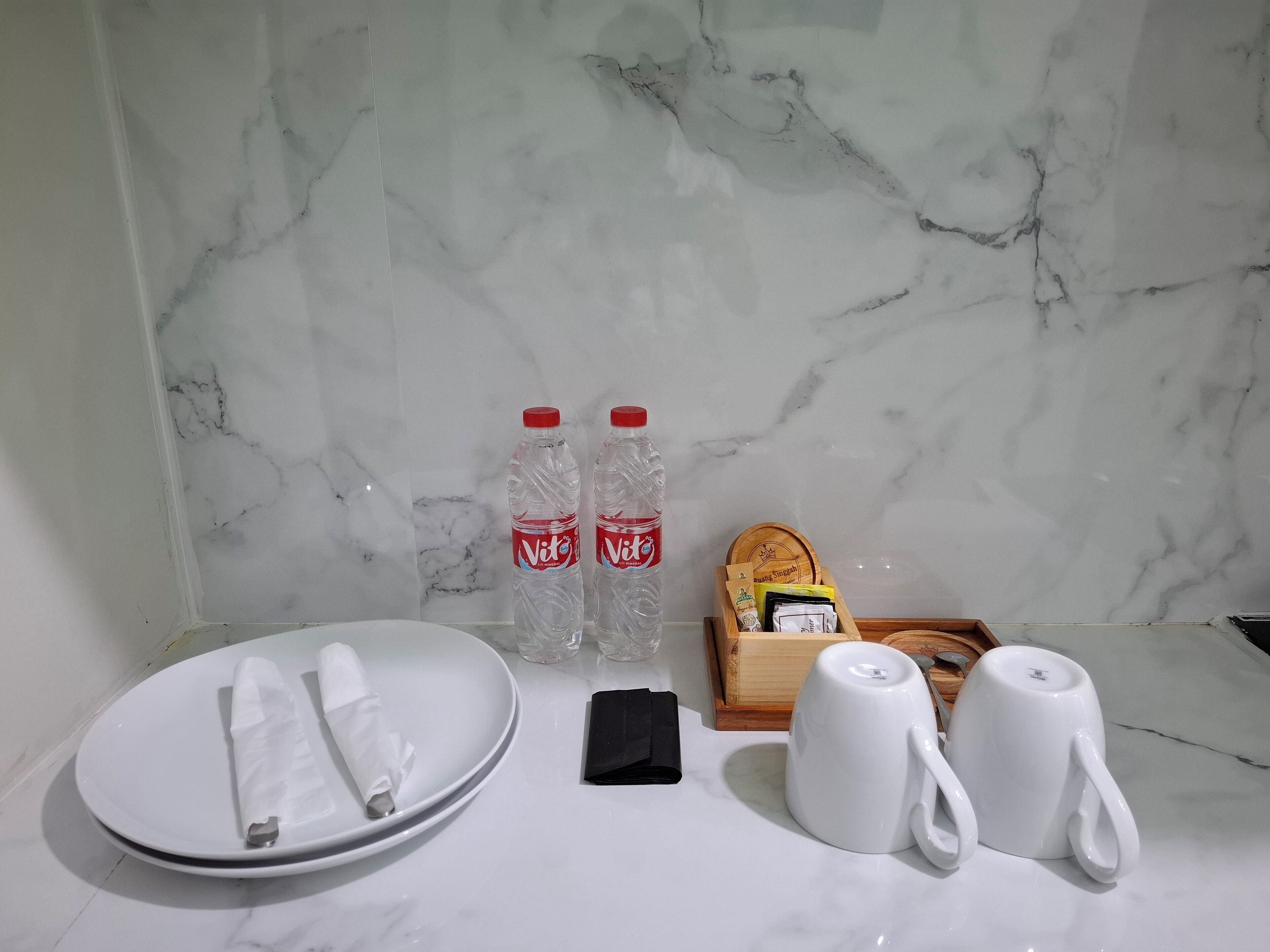 A bathroom with a marble wall, a white sink, two water bottles, a toothbrush, a cup, and a box of tissues.