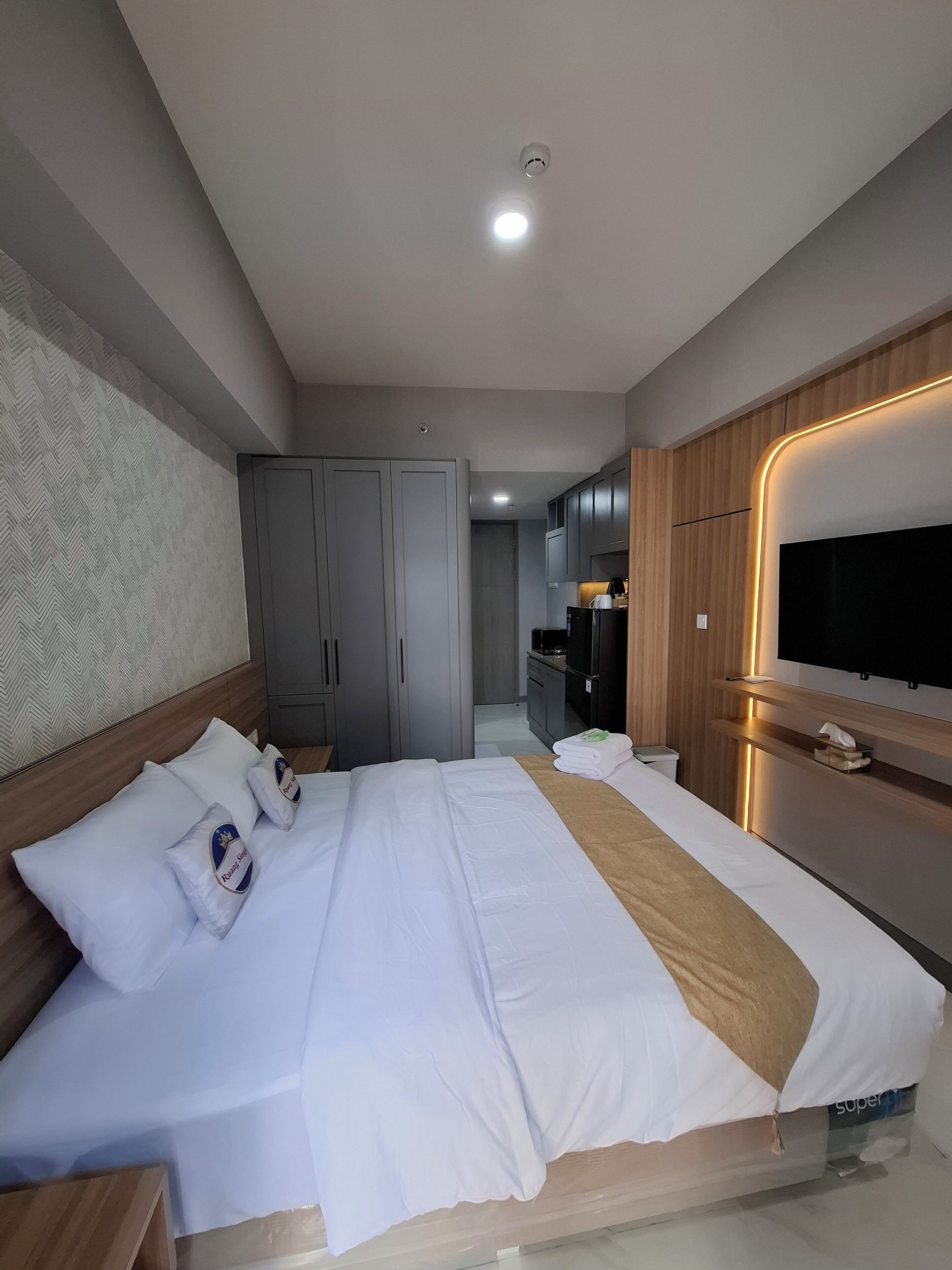 A modern hotel room with a large bed, a TV mounted on the wall, and a kitchenette in the background.