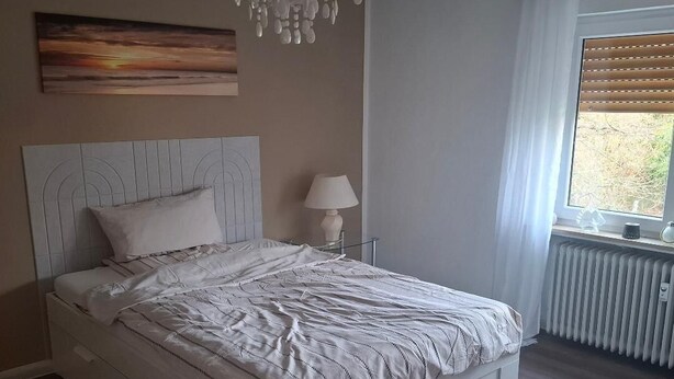 Une chambre à coucher avec un lit blanc, une table de chevet, une lampe, un tableau au mur, une fenêtre avec des stores et un radiateur.