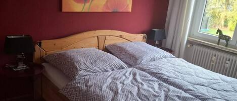 Un dormitorio con cama de madera, ropa de cama estampada, pared roja, cuadro de flores, mesita de noche, lámpara y ventana con vistas.