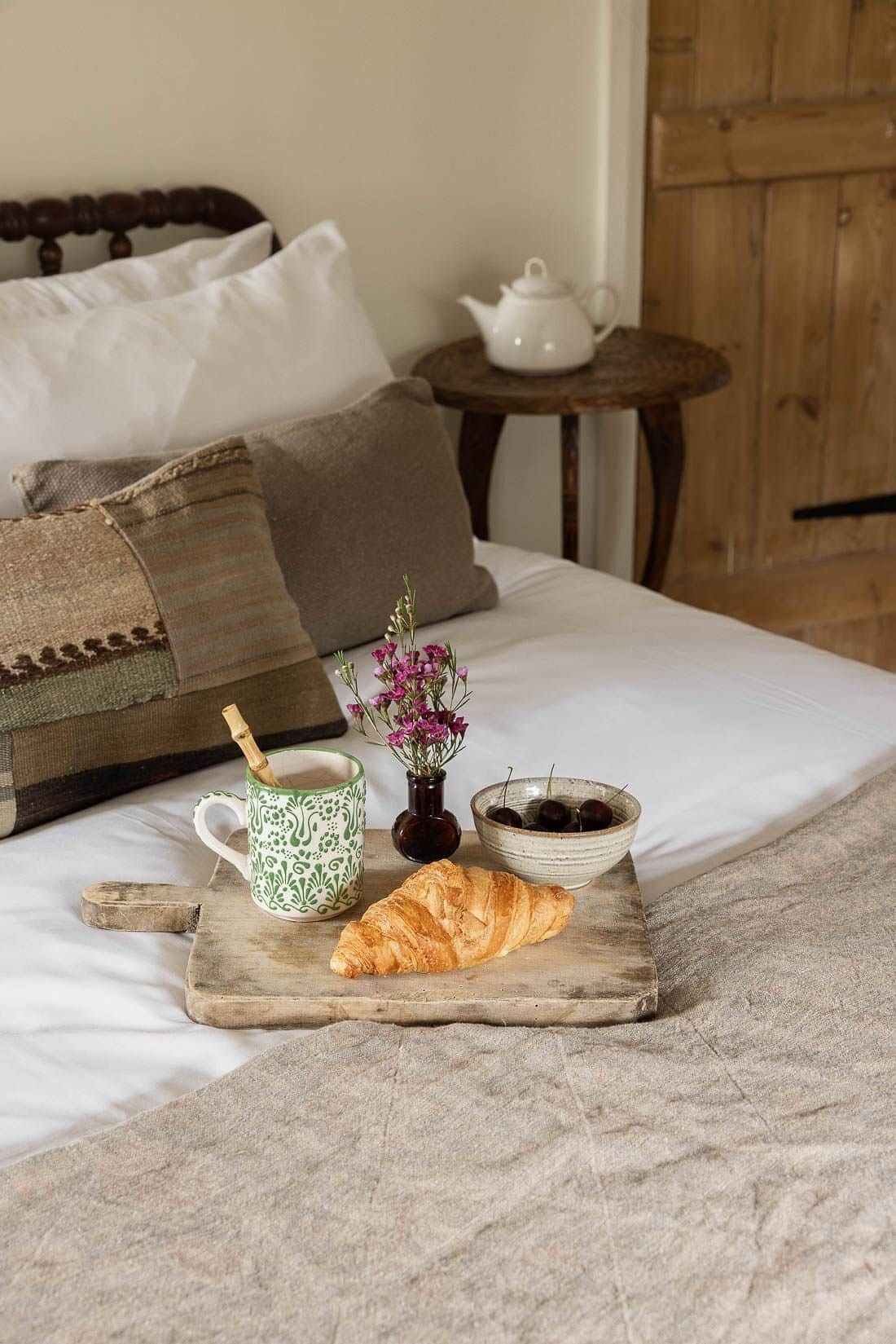A bed with pillows, a bedside table with a teapot, a tray with a croissant, a mug, a bowl of cherries, and a vase of flowers.