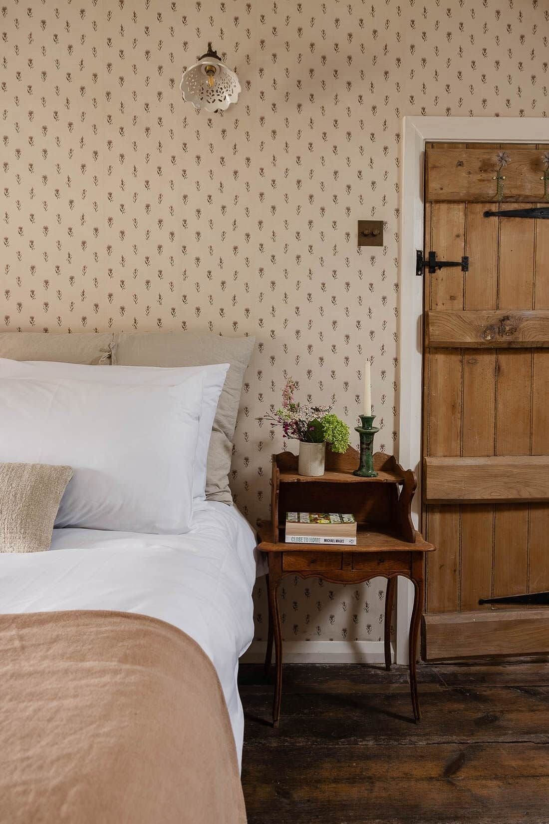 A bedroom with a bed, a bedside table with a book, a wall-mounted light, and a wooden door.