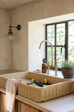 A kitchen with a large sink, a window with a view of greenery, a hanging light fixture, and a potted plant on the windowsill. - The Curated Country House (Chipping Campden)