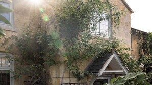 Un bâtiment en pierre avec une porte en bois, un petit jardin avec différentes plantes et un ciel dégagé.