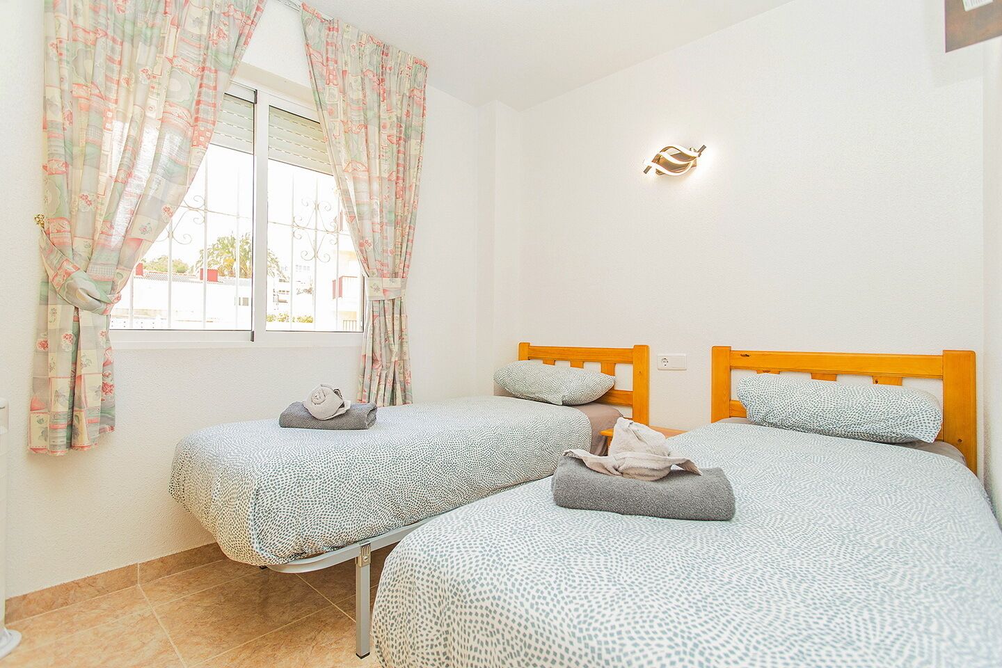 A bedroom with two beds, a window with curtains, a wall-mounted light, and a tiled floor.