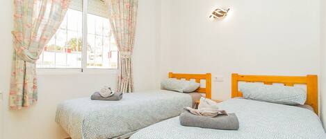A bedroom with two beds, a window with curtains, a wall-mounted light, and a tiled floor.