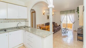 Eine Küche mit weißen Schränken, einer Granit-Arbeitsplatte und einem Spülbecken. Es gibt einen Essbereich mit Tisch und Stühlen, sowie eine Couch.
