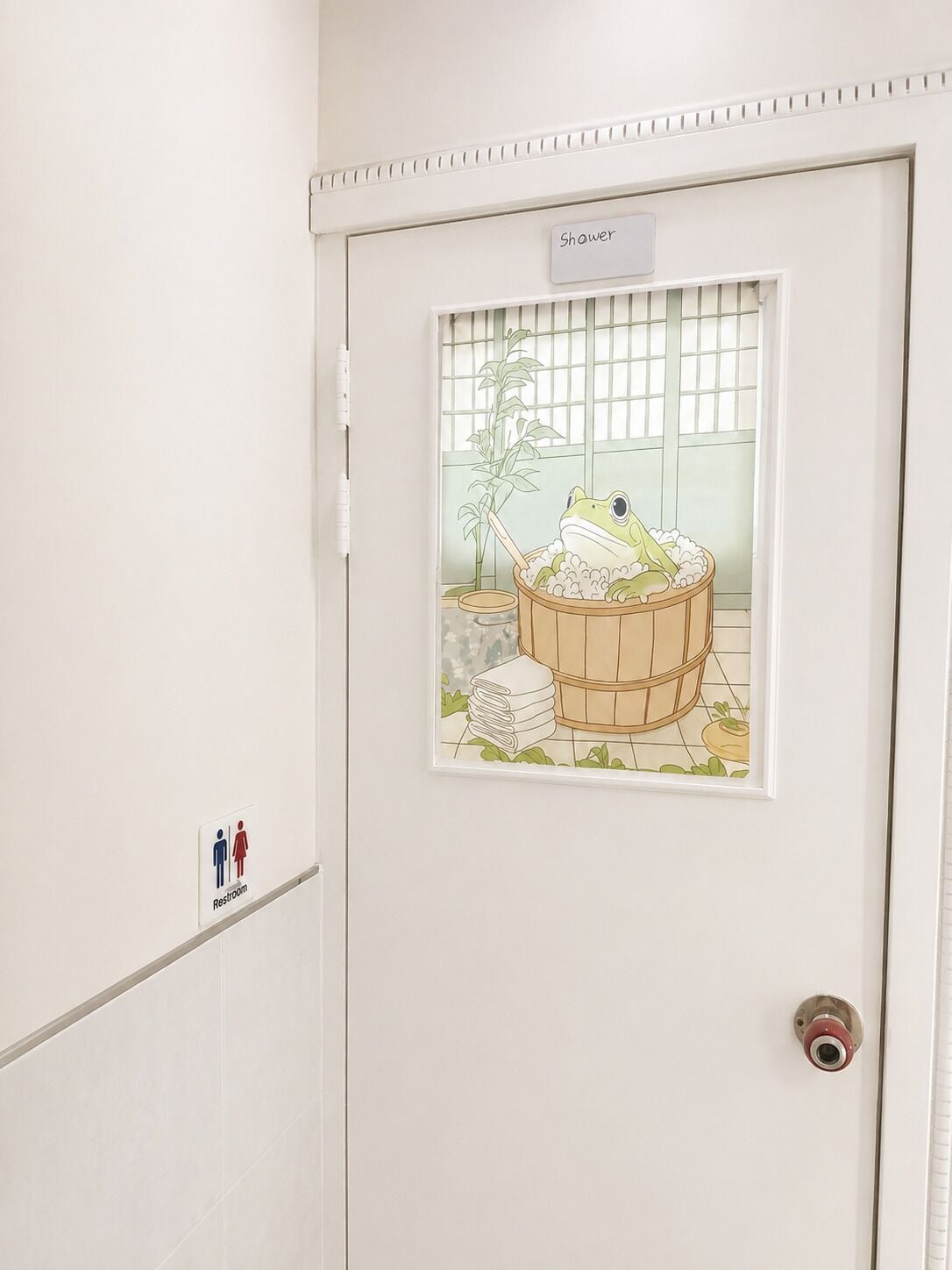 Une porte de salle de bain avec une fenêtre en verre dépoli représentant un dessin animé d’une grenouille dans une baignoire, un panneau indiquant la présence d’une douche et un panneau indiquant les toilettes.