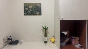 Une pièce avec un mur blanc, un tableau représentant des fleurs, une bouteille de vin, une petite plante et un jouet en peluche.