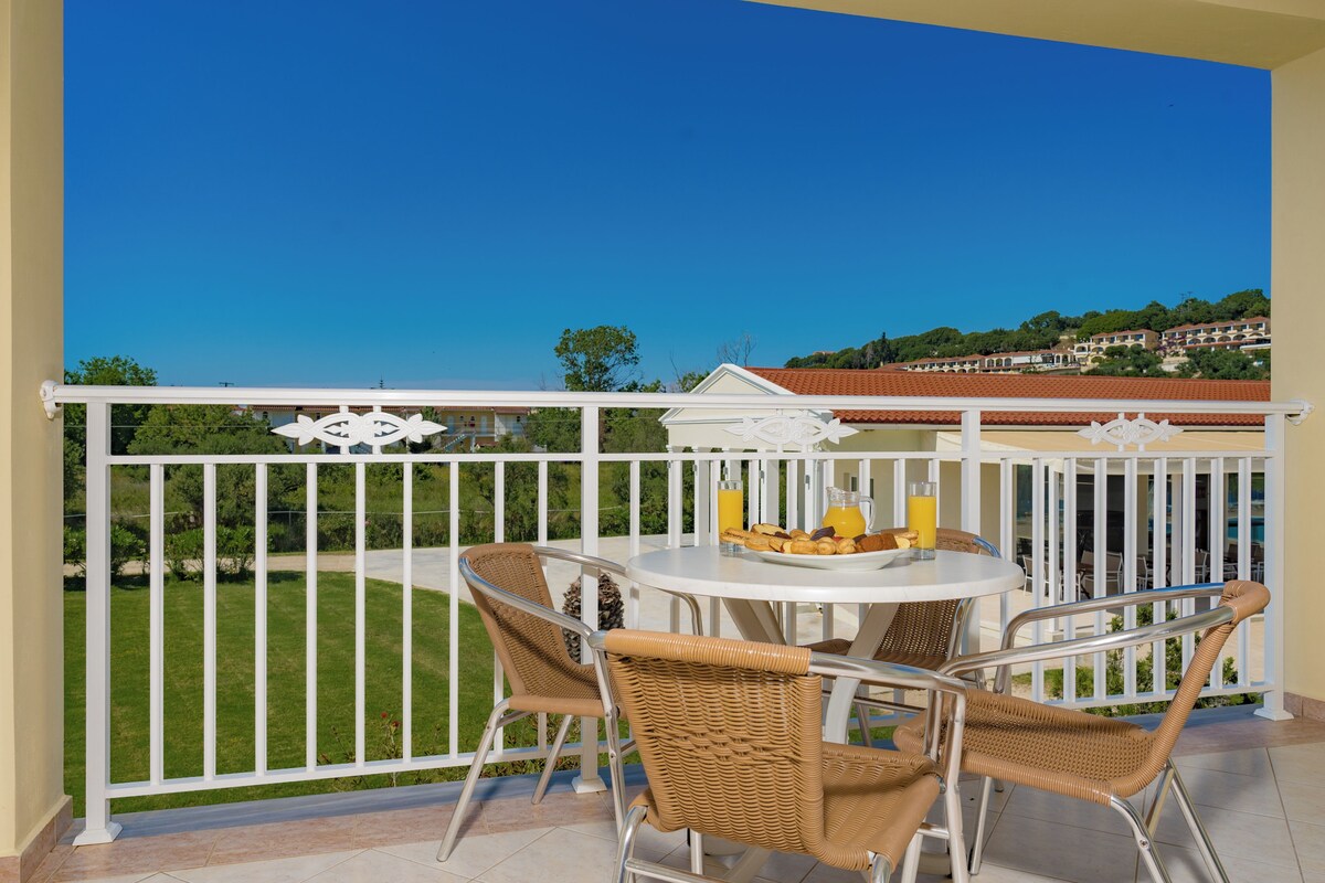 A balcony with a white railing, wicker chairs, a round table with a fruit plate and a glass of orange juice, and a view of a green landscape and buildings in the background.